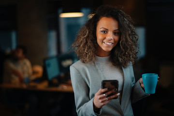 Young businesswoman using phone and drinking coffee in office.	