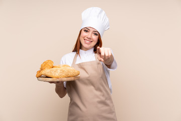 Redhead teenager girl in chef uniform. Female baker holding a table with several breads points finger at you with a confident expression