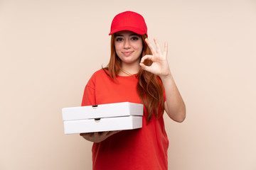 Pizza delivery teenager girl holding a pizza over isolated background showing an ok sign with fingers