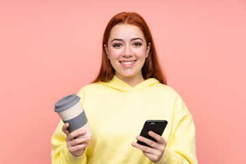 Fototapeta premium Redhead teenager girl over isolated pink background holding coffee to take away and a mobile
