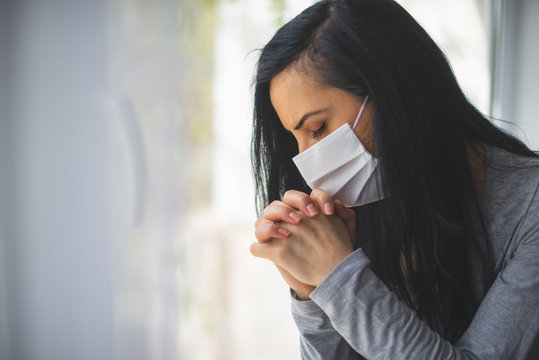 Portrait Of Woman With Surgical Mask Praying Next To Window