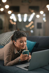 Young woman listening music. Beautiful woman sitting on sofa and drinking coffee in living room.	