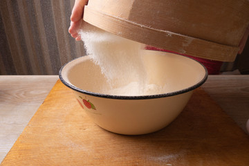 Large bowl, cutting board, sieve for sifting flour on the table. Female hands sifting flour in the home kitchen.