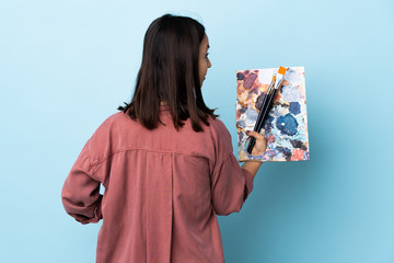 Young artist woman holding a palette over isolated blue background in back position and looking side.