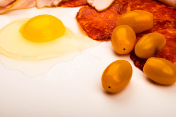 A broken chicken egg without a shell, slices of sausage and bacon and tomatoes on a white background. Close up.