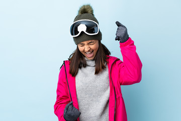 Mixed race skier girl with snowboarding glasses over isolated blue background celebrating a victory.