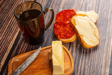 Fresh wheat toast with butter, sausage and cheese, a wooden butter dish with a piece of butter and a mug of tea on a wooden background. Close up.