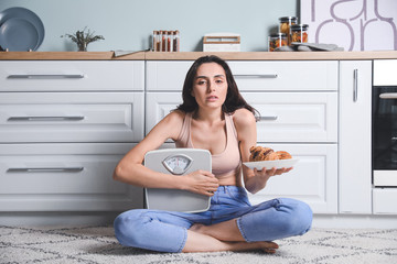 Young woman suffering from anorexia with croissants and scales in kitchen