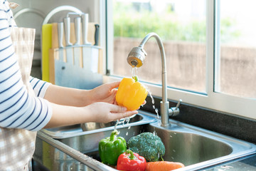 Asian healthy woman washing yellow bell pepper and other vegetable above kitchen sink and cleaning...