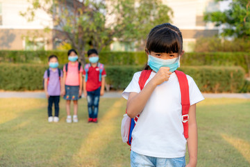 Asian girl preschool child student wearing healthy face mask sneeze with friends in background. WHO recommends at least 6 feets of distance between yourself and people who are coughing or sneezing.