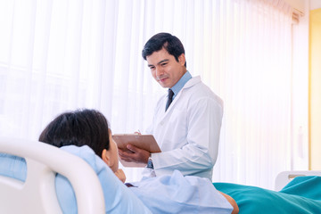 Obraz premium Hospitalized young woman lying in bed while doctor checking her healthcare. Friendly man physician examining female patient in hospital room ward while holding clipboard report in his hand.