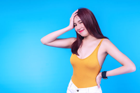 Cheerful Young Woman Brown Long Hair Wear Short Jeans While Standing On Isolated Blue Background. Positive Teenager Girl Smiling Posture While Looking At Camera With Copy Space.