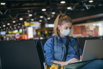 A woman is sitting at the airport with a laptop in a medical mask