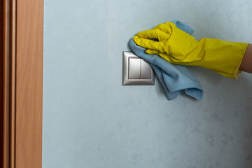 Hand in protective yellow gloves is cleaning the light switch. Disinfecting surfaces in the house during COVID19 quarantine.