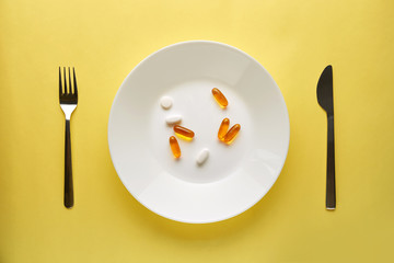 Pharmaceutical medicine pills or capsules on a plate with a fork and knife. Top view yellow background.