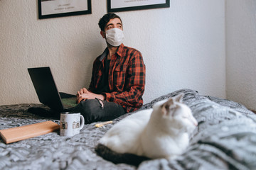 A young man with medical mask works on a laptop in his bed with his cat in quarantine time