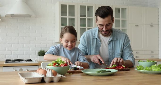 Happy cute small kid daughter cooking with father looking at camera stand at kitchen table. Young dad teaching child learning preparing healthy food cutting vegetable salad. Vegan family making meal.