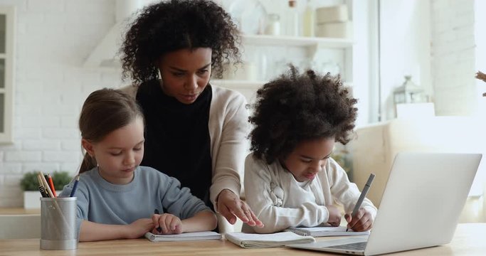 Happy young adult african mom, nanny, tutor helping two mixed race cute elementary school girls studying together doing homework. Diverse ethnicity children home education, biracial family parenting.