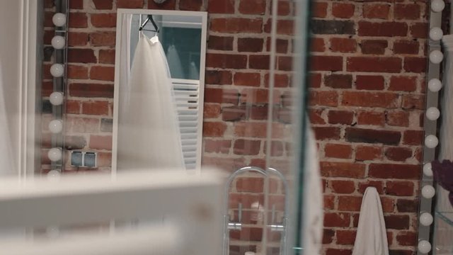 Hanging wedding outfit for bride on the glass door in front of brick wall and reflection in the mirror of the part of wedding clothing