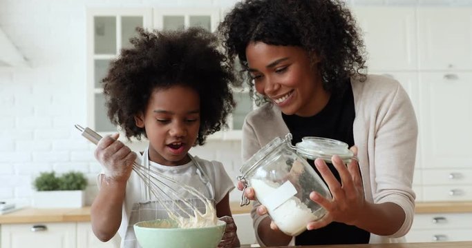 African mom and small kid daughter cooking together making dough in bowl in kitchen. Happy mixed race family prepare cake mix ingredients for pie having fun baking pastry on holiday morning at home.