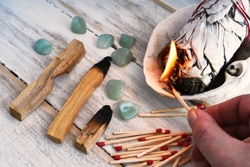 A close up image of a hand holding a lit match to a white sage smudge stick. 