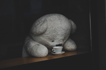 A fluffy, white depressed teddy bear hunched over an empty cup of coffee at closed cafe. 