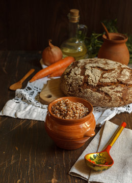 Buckwheat porridge in a pot and a wooden painted spoon on a linen napkin, black bread and vegetables in the background