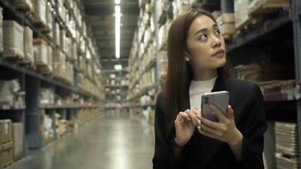 Asian female worker wearing a black suit Checking in warehouse For her company To export to customers By using a smartphone to take notes.