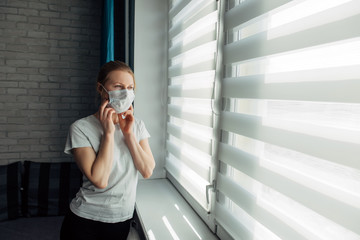 Young woman in mask looking through the blinds. Quarantined coronavirus person. Stay home. Stop the pandemic. Self-isolation, putting up mask.