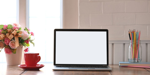 Photo of computer laptop with white blank screen putting on wooden working desk and surrounded by notebook, bunch of flowers, cup and pencil holder over comfortable living room windows as background.
