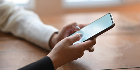 Cropped of woman's hands holding a white blank screen smartphone while sitting and relaxing at the wooden working desk over comfortable living room windows as background.