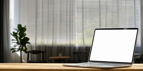 Fototapeta premium Photo of Computer laptop with white blank screen putting on wooden working desk over living room curtain and potted plant as living room background.