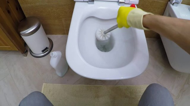 POV of man in rubber gloves using stiff-bristled brush and cleaning toilet bowl at home