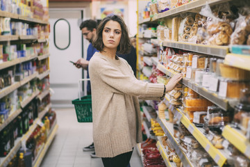 Pensive female buyer in grocery store. Thoughtful young woman standing near shelves and choosing goods in supermarket. Shopping concept
