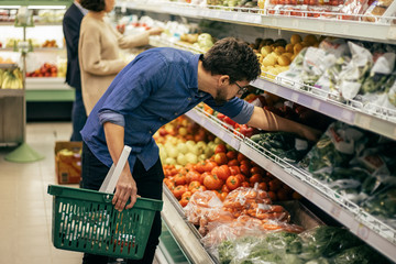 Focused people shopping in grocery store. Side view of men and woman holding shopping baskets and choosing fresh fruits and vegetables in supermarket. Shopping concept