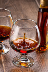 two glasses with cognac and splash next to a bottle on a wooden background