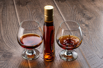 two glasses with cognac and splash next to a bottle on a wooden background