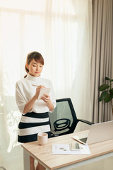 Young Asian woman writing notebook while standing behind working desk at home