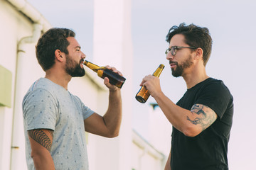 Two handsome young man drinking beer. Smiling friends with beer bottles hanging out on balcony....