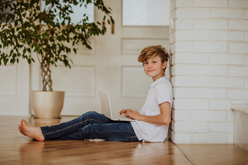 Young  blond boy in white shirt and jeans printing on the white laptop sitting on the floor at home © Alina