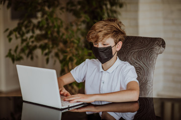 Blond cute young boy in in white shirt and black medical mask printing on the white laptop at home