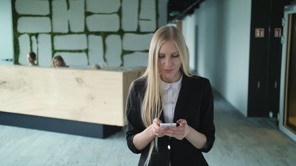 Stylish blond woman in jacket standing in contemporary office hall and surfing smartphone with smile