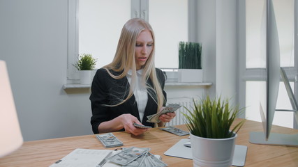Beautiful young blond woman in business suit, sitting in office at light wooden desk with computer concentrating on counting large pile of cash in hands