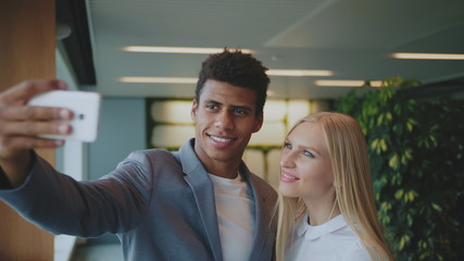 Fototapeta premium Cheerful black man with laughing blond woman taking selfie with smartphone in modern office having fun.