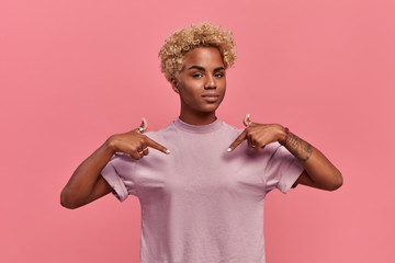 Self assured proud African American woman has blonde hairstyle satisfied with her own high achievements, points with two hands at herself, or at a t-shirt for printing, isolated over pink background