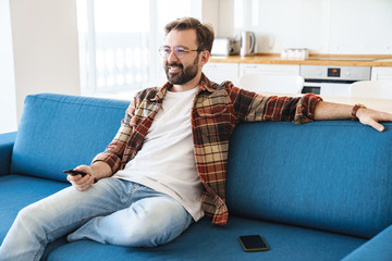 Obraz premium Portrait of young bearded man smiling and watching television at home