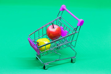 Self-service shopping cart with pink handle and mini toy fruits isolated on green background....