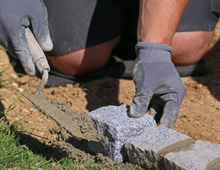 Close up photo of worker with trowel and gloves, laying a lawn edge massive granite blocks as a...