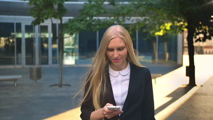 Fototapeta premium Content blond woman in black jacket holding smartphone and looking away with smile on street background