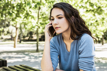 Calm young woman sitting on bench and talking on phone. Tranquil lady listening interlocutor through phone while sitting in park. Technology and communication concept